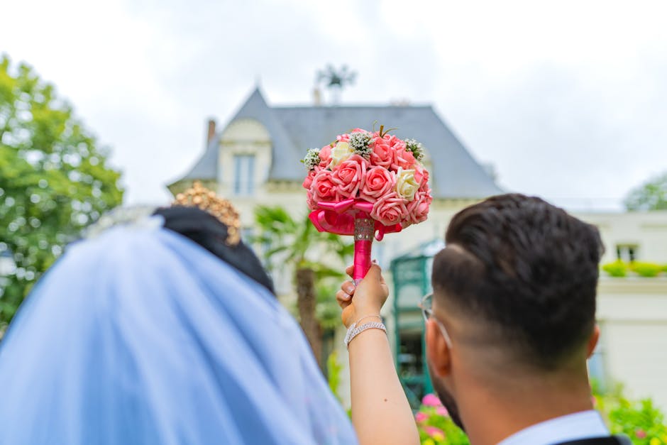 Close-up of a bouquet arrangement held by a person during a wedding, featuring pink and white roses with some small white filler flowers. The bouquet is wrapped in bright pink paper and ribbon. The pe