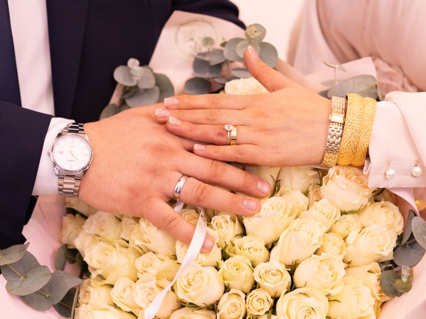 Close-up of a bouquet arrangement held by a person during a wedding, featuring pink and white roses with some small white filler flowers. The bouquet is wrapped in bright pink paper and ribbon. The pe