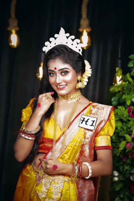 A young woman dressed in traditional Indian attire, wearing a vibrant yellow saree with intricate gold embroidery and a contrasting maroon border, is adorned with elaborate gold jewelry including a ch