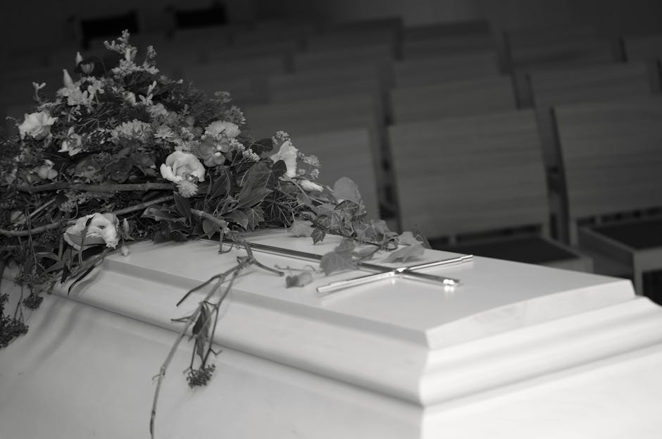 A floral arrangement placed on a wooden surface featuring a mix of white and cream-colored flowers, including hydrangeas and roses, accompanied by lush green leaves. Behind the bouquet, tall, dried be